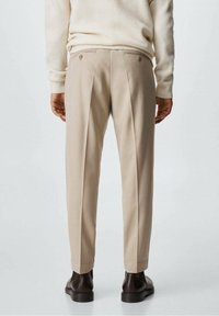 Pantaloni beige a gamba dritta con due bottoni posteriori, una parte anteriore plissettata e una texture liscia. Indossati con un maglione crema chiaro e stivali scuri.