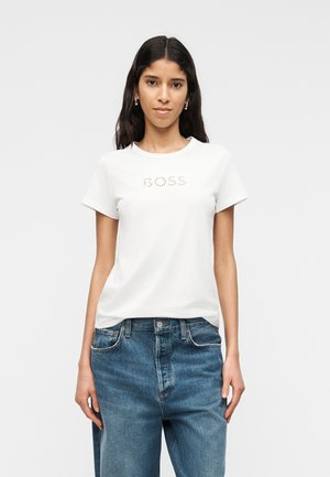 Kobieta z długimi ciemnymi włosami, ubrana w białą koszulkę z napisem "BOSS" i luźne niebieskie dżinsy, stojąca na tle prostego, jasnego tła.