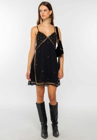 Robe sans manches noire avec des embellissements en étoiles dorées, présentant un design triangulaire et un ourlet transparent. Portée avec des bottes noires jusqu'aux genoux.