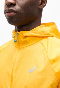 Κοντινό πλάνο ατόμου που φοράει κίτρινο μπουφάν με κουκούλα, με μπλε φερμουάρ και λευκό λογότυπο στο στήθος, πάνω σε λευκό φόντο.