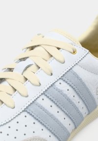 Άσπρο δερμάτινο αθλητικό παπούτσι με γαλάζιες ρίγες, κρέμ κορδόνια, χρυσές τρύπες και τρύπες κατά μήκος της πλευράς. Υφασματικά σουέντ διακοσμητικά στοιχεία.