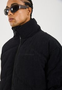 Veste zippée en velours côtelé noir avec un col montant, un design matelassé horizontal et une fermeture éclair à l'avant. Présente un logo discret sur la poitrine.
