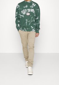 Sweatshirt vert avec des motifs de feuilles blanches et le texte "PLACE IN THE WOODS", associé à un pantalon beige et des baskets blanches.