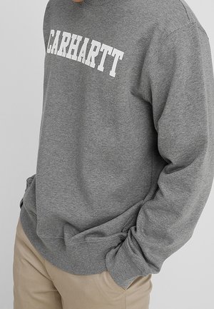 Person trägt ein graues Carhartt-Sweatshirt und beige Hosen, mit einer Hand in der Tasche, vor einem schlichten hellen Hintergrund.