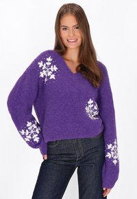 Maglione lavorato a maglia viola con scollo a V, caratterizzato da motivi di fiocchi di neve bianchi sulle maniche. Texture morbida e vestibilità ampia. Indossato con jeans scuri.