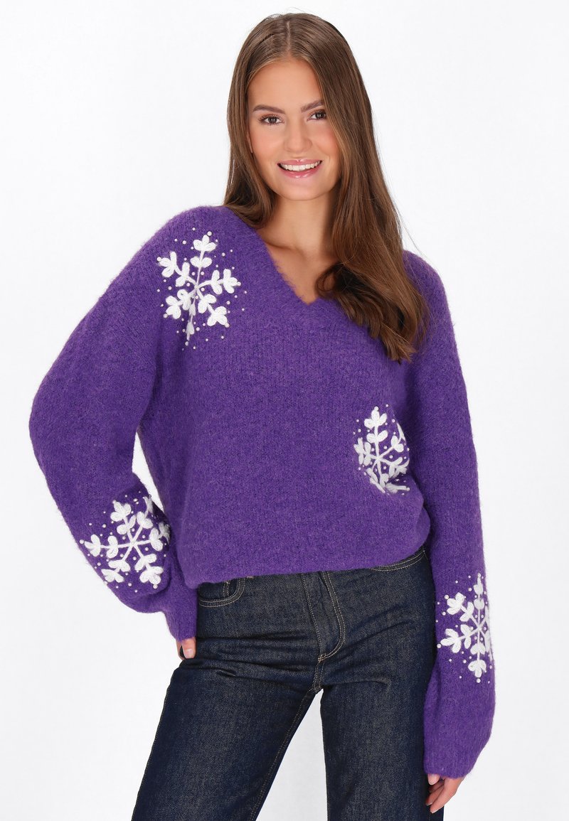 Maglione lavorato a maglia viola con scollo a V, caratterizzato da motivi di fiocchi di neve bianchi sulle maniche. Texture morbida e vestibilità ampia. Indossato con jeans scuri.
