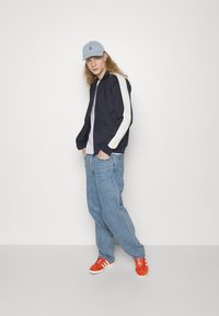 Veste de bombardier marine avec des rayures blanches, t-shirt blanc, casquette bleu clair, jeans en denim bleu et baskets orange avec des accents blancs.
