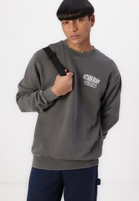 Gray sweatshirt with round neckline, ribbed cuffs, and hem, featuring a white logo on the chest. Model holds a black shoulder strap.