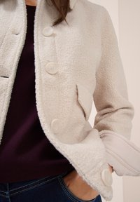 Beige strukturerad knäppjacka över en mörk lila tröja, hand i fickan på blå denimjeans, närbild på bål och arm.