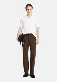 Polo bianco, pantaloni marroni, cintura nera e calzature. Tiene una borsa nera con una finitura liscia. Silhouette semplice e su misura.