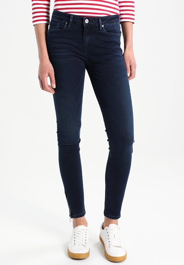 Person trägt dunkelblaue Skinny-Jeans mit Reißverschlüssen am Knöchel, weiße Sneakers mit Gummisohlen und ein rot-weiß gestreiftes Langarmshirt.