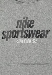 Pelēks kapucis, izgatavots no flīsa auduma, ar lielu melnu "nike sportswear" logo un "ESTABLISHED 1972" baltā tekstā.