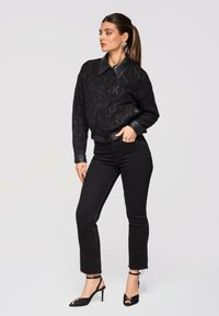 Femme debout, main dans la poche, portant une veste matelassée noire avec un col et des poignets en cuir, un jean noir et des talons noirs à brides.