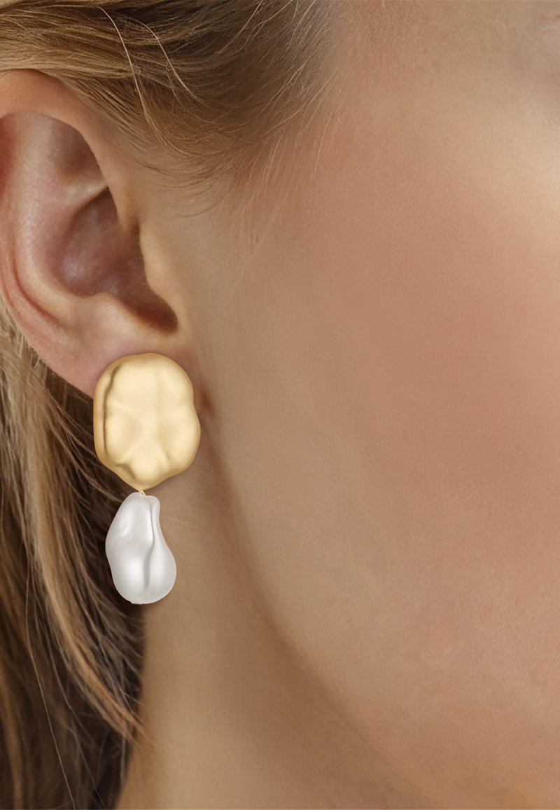 Primer plano de la oreja de una mujer que lleva un pendiente desparejado con una pieza superior de oro texturizado y una gota blanca de forma irregular.