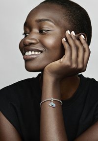 Strieborný náramok so súprava dvoch farebných príveskov. Náramok má textúrovaný povrch a je nosený na zápästí s tmavým tričkom.