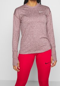 Maglia sportiva bordeaux a maniche lunghe con scollo tondo e fori per il pollice, abbinata a leggings rossi con logo Nike Swoosh nero.