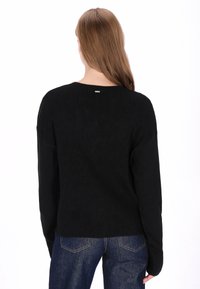 Fekete kötött pulóver bordázott mintával, laza szabással és egy kis díszítéssel a nyakkivágás közelében. Kék farmernadrággal párosítva.