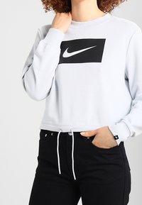 Světle šedý zkrácený svetr s černým obdélníkovým logem. Má dlouhé rukávy, kulatý výstřih a nastavitelnou šňůrku na spodním lemu.