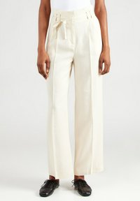 Crème brede lange broek met een strik tailleband, geplooid voorpand en twee zijzakken. Gepaard met zwarte veterschoenen.