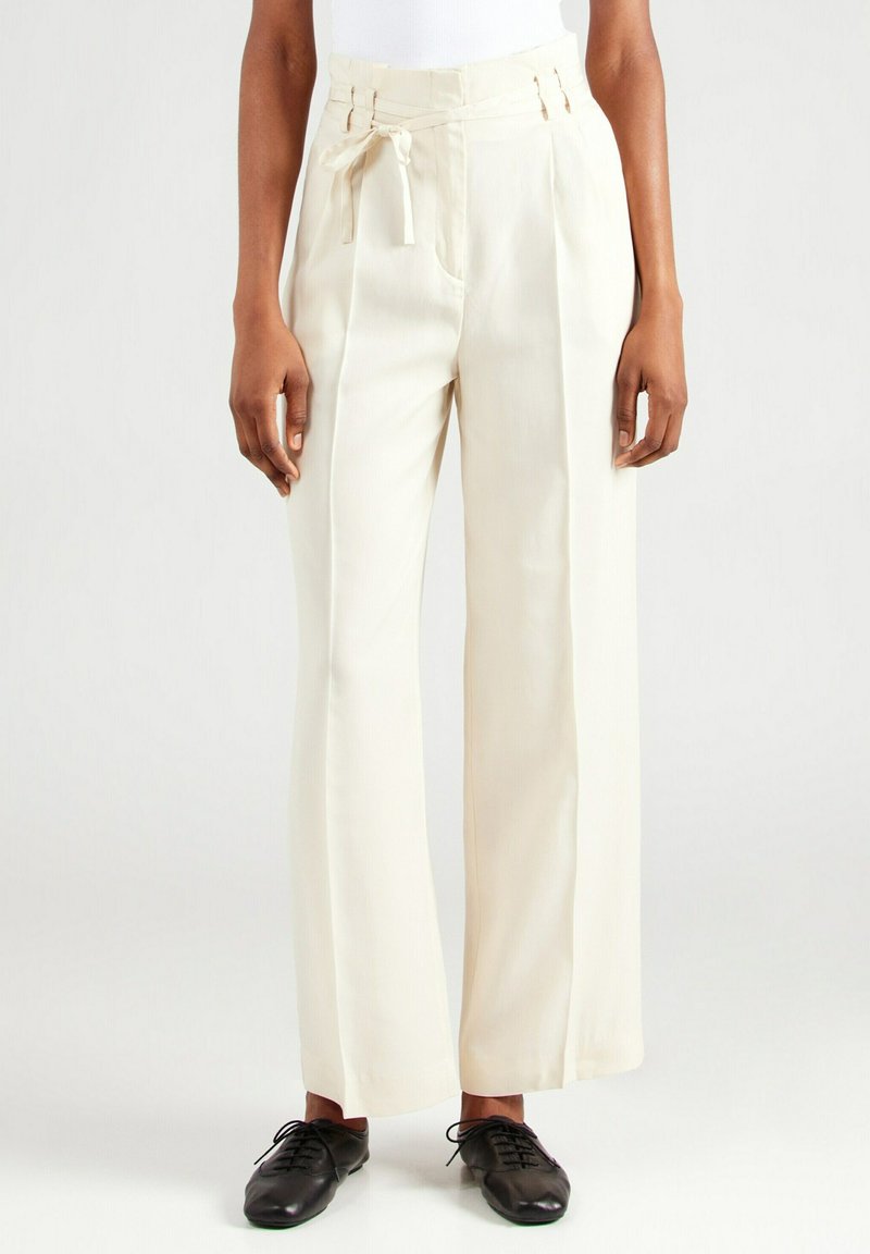 Crème brede lange broek met een strik tailleband, geplooid voorpand en twee zijzakken. Gepaard met zwarte veterschoenen.