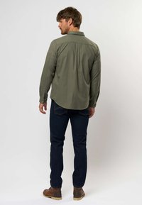 Camicia verde a maniche lunghe con colletto a punta, polsini con bottoni e dettaglio con piega sul retro; abbinata a jeans scuri e calzature marroni.