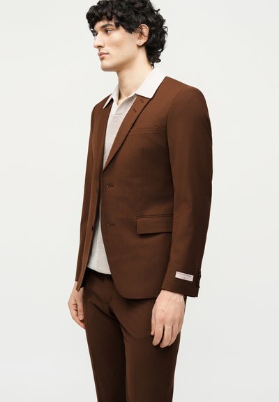 Άνδρας με σγουρά μαλλιά, ντυμένος με καφέ κοστούμι και ανοιχτό μπεζ πουκάμισο με γιακά, στέκεται προφίλ μπροστά σε ένα απλό ανοιχτό φόντο.