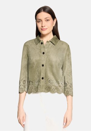 Vrouw draagt een olijfgroene jas met knopen en een bloemenuitsnede aan de zoom, met driekwartmouwen, gecombineerd met een witte broek.