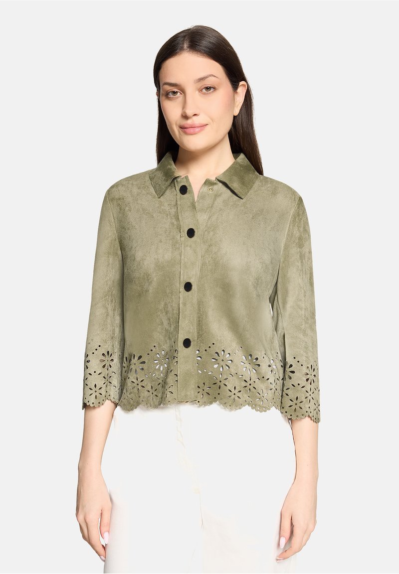 Femme portant une veste boutonnée vert olive avec un ourlet découpé à motif floral et des manches trois-quarts, associée à un pantalon blanc.