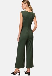 Green jumpsuit with a sleeveless design, a V-back, and wide legs, made from a smooth fabric. Paired with black block-heel sandals.