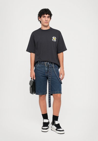 Jeune homme portant un T-shirt graphique noir, des shorts en denim avec une ceinture à œillets longue, des chaussettes noires, des baskets noir et blanc, et tenant un sac à main noir.