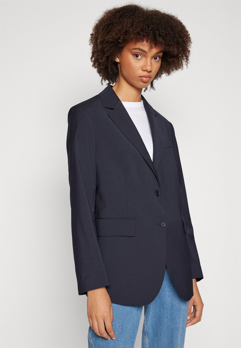 Blazer marine à fermeture à un seul bouton, avec des revers crantés et deux poches avant, fabriqué dans un tissu lisse. Modèle associé à un jean bleu clair.