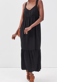 Robe maxi noire avec un col en V, des bretelles épaisses, une taille froncée et un ourlet à volants. Associée à des sandales marron à brides et un sac à bandoulière marron.