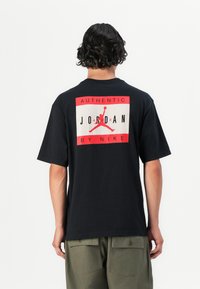 Sort kortærmet t-shirt med et stort rød- og hvidt grafisk motiv på ryggen, der viser "AUTHENTIC JORDAN BY NIKE" og en springende mand-silhuet.