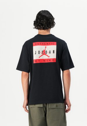 T-shirt noir à manches courtes avec un grand motif rouge et blanc dans le dos, avec l'inscription "AUTHENTIC JORDAN BY NIKE" et la silhouette d'un homme sautant.