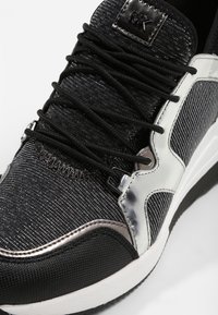 Zapatilla deportiva negra y plateada con tejido texturizado, cordones negros, suela blanca y logo "MK" en la lengüeta.
