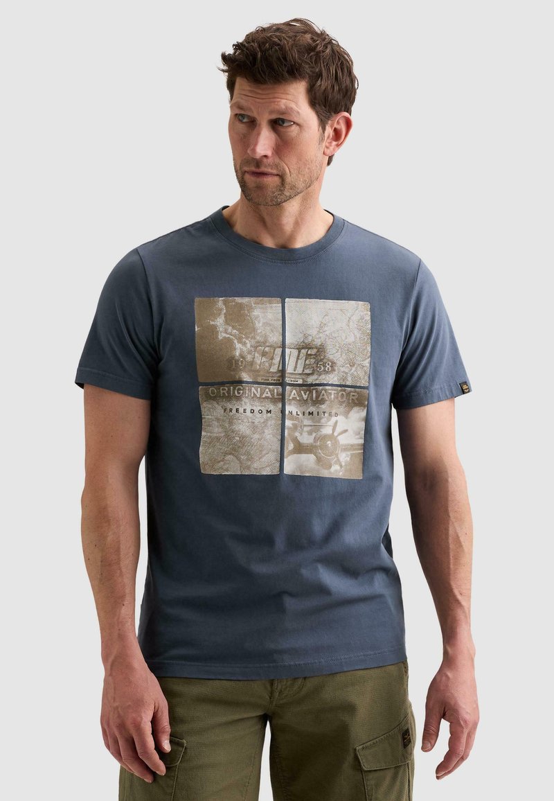 Άνδρας που φοράει μπλε γραφικό T-shirt με θέμα τον αεροπόρο και ελιάς πράσινο παντελόνι cargo, κοιτάζει προς τα πλάγια.