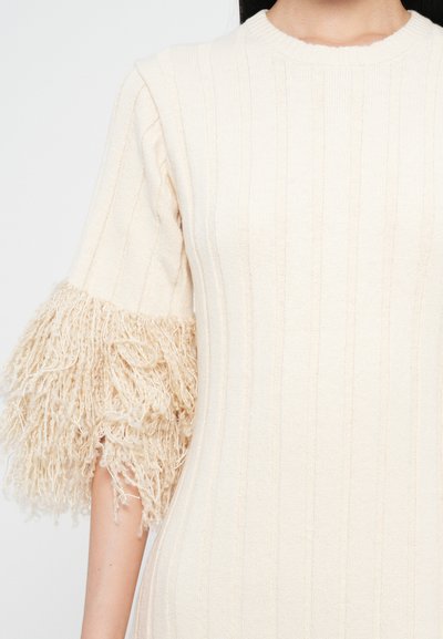 Robe tricotée en blanc cassé, présentant une texture côtelée et des manches courtes avec de doux accents de franges beiges au niveau des poignets. Encolure ronde simple.