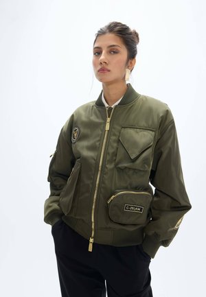 Joven mujer con el pelo recogido lleva una chaqueta bomber verde con múltiples bolsillos y parches, de pie frente a un fondo liso y claro.