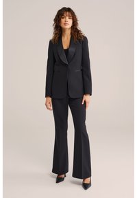 Kvinna med lockigt hår som bär en svart skräddarsydd kavaj, utsvängda byxor och svarta spetsiga klackar, stående mot en enfärgad bakgrund.