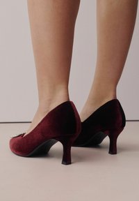 Piernas con zapatos de tacón medio de terciopelo rojo oscuro con un detalle de piedras brillantes en la parte delantera, sobre un suelo de color claro.