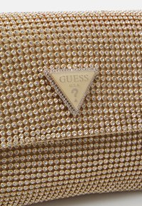 Zlatna torbica s kopčom prekrivena malim, okruglim rhinestones; sadrži trokutasti logo u sredini s imenom marke "GUESS" i upitnikom.