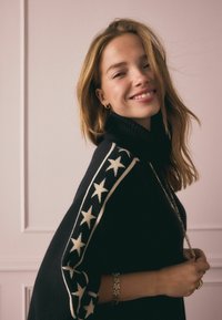 Fekete kötött garbó fehér csillagminta a ujjak mentén. A modell arany karkötőt és medálos nyakláncot visel, halvány háttér előtt.