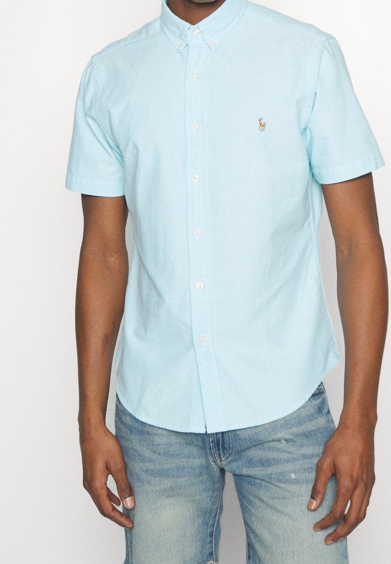 Světle modrá košile s krátkým rukávem a knoflíkovým zapínáním, vyrobená z bavlny, s límečkem zapínaným knoflíky a malým vyšitým logem na levé hrudi.