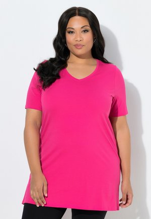 Frau mit langen dunklen Haaren, die eine leuchtend pinke Kurzarm-Tunika über schwarzen Leggings trägt und vor einem schlichten weißen Hintergrund steht.