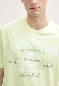 Lys gul T-shirt med rund halsudskæring, der har hvid og grøn trykt tekst samt et cirkulært grafisk design. Blød tekstur, afslappet pasform.