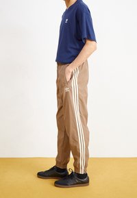 Person som står sidledes och bär bruna Adidas trackbyxor med vita ränder, en blå Adidas t-shirt, svarta strumpor och svarta Adidas Samba skor.