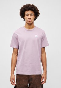 Απαλό μωβ βαμβακερό t-shirt με κοντά μανίκια, στρογγυλή λαιμόκοψη και μια μικρή μπλε ετικέτα στο στήθος. Λείο υλικό, άνετη εφαρμογή.