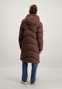 Bruine gewatteerde jas met capuchon, voorzien van horizontale gestikte textuur en rits sluiting. Gepaard met blauwe spijkerbroek en witte sneakers.