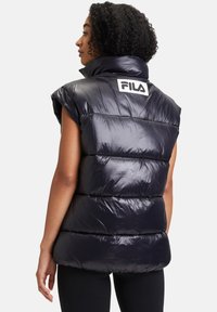 Puffa väst i glänsande marinblått material med hög krage, korta ärmar och horisontellt quiltade sektioner. Har en vit FILA-logga på baksidan.