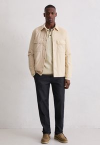 Beżowa kurtka z dwoma kieszeniami na piersi, kremowy sweter, ciemne spodnie i beżowe buty. Gładka tkanina, luźny krój, warstwowy wygląd.